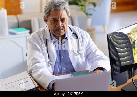 Serious Senior biracial männlichen Arzt sitzt am Schreibtisch mit Laptop Stockfoto