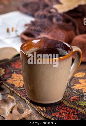 Heiße Tasse Kaffee auf der Herbstmatte mit Buch und Decke im Hintergrund Stockfoto