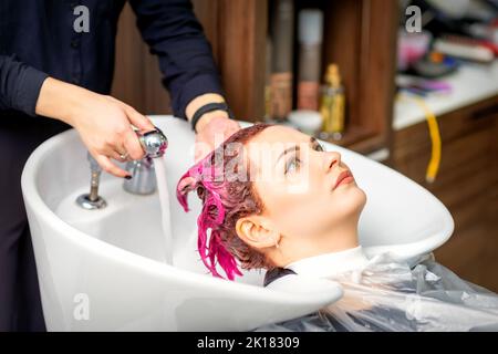 Waschen gefärbte weibliche Haare. Eine junge kaukasische Frau, die in einem Schönheitssalon ihre Haare waschen ließ. Der professionelle Friseur wäscht die rosa Farbe von den Haaren eines Kunden ab Stockfoto