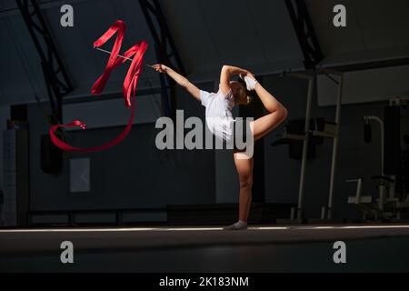 Flexibles kleines Mädchen, weibliches rhythmisches Turnertraining mit rotem Band in der Sporthalle, drinnen. Konzept von Aktion, Bewegung, Sport, Motivation Stockfoto