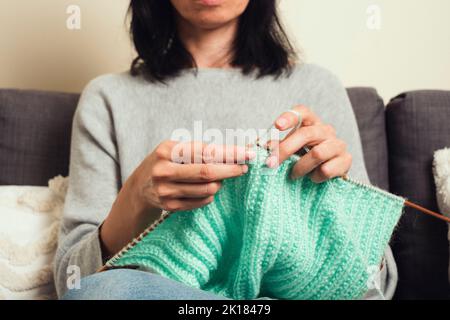 Nahaufnahme der weiblichen Hände, die Bambusstricknadeln halten und grünen Wollpullover stricken. Hobby, Entspannung, psychische Gesundheit, nachhaltiger Lebensstil Stockfoto