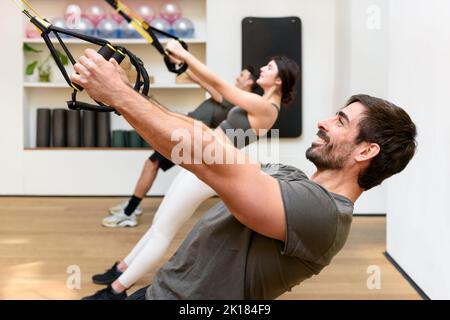 Seitenansicht des bärtigen männlichen Athleten, der lächelt und während des Gruppentrainings im Fitnessstudio mit TRX-Seilen in niedrigen Reihen trainiert Stockfoto