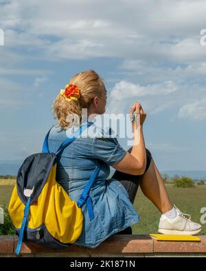 Die blonde Frau mit Rucksack, die auf einer niedrigen Wand sitzt, gibt vor, auf dem Himmel vor ihr zu zeichnen Stockfoto