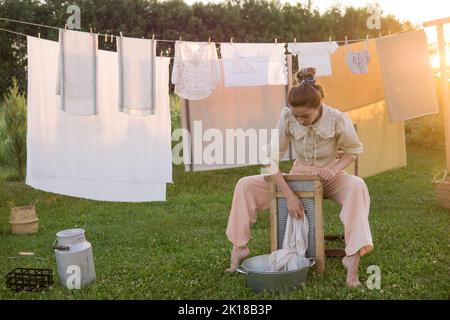 Wäschereitag. Eine Frau hängt Bettwäsche und Handtücher auf einem Baum im Hof eines Dorfhauses. Sommerhaus-Konzept. Stockfoto
