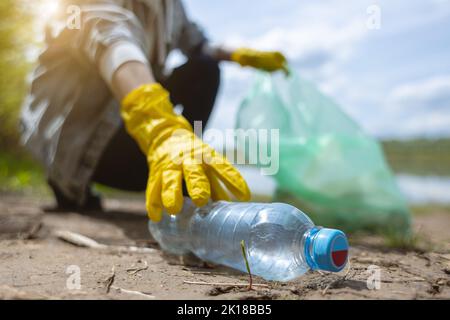 Eine junge Frau in gelben Handschuhen sammelt verlassenen Müll in einer schwarzen Tasche im Wald. Verschmutzung durch Plastik und Umweltschutz Stockfoto