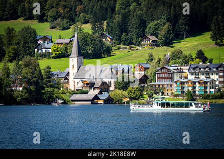 Blick auf ein Ausflugsboot, das auf dem Altausseer See fährt, vorbei an der historischen Stadt Altaussee, dem Ausseer Land, der Steiermark, Österreich und seiner katholischen Kirche Stockfoto