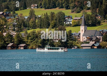 Blick auf ein Ausflugsboot, das auf dem Altausseer See fährt, vorbei an der historischen Stadt Altaussee, dem Ausseer Land, der Steiermark, Österreich und seiner katholischen Kirche Stockfoto