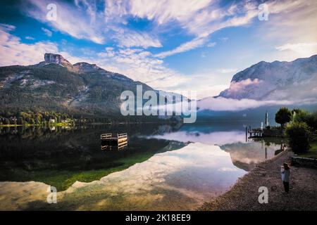 Atemberaubende Morgenansicht einer Frau, die am Ufer des Altausseer Sees, der Steiermark, Österreich, beim Kaffeetrinken und beim Blick auf den See steht Stockfoto