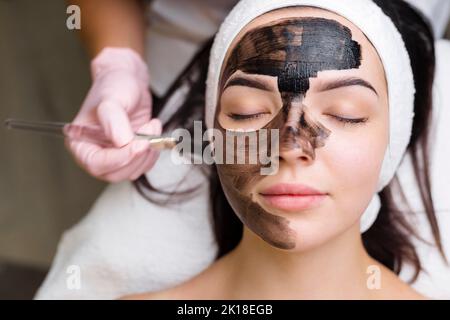 Kosmetologe, der schwarze Maske auf hübsches Frauengesicht mit schwarzen Handschuhen aufträgt, wunderschöne Frau im Spa, die Gesichtsbehandlungen hat Stockfoto