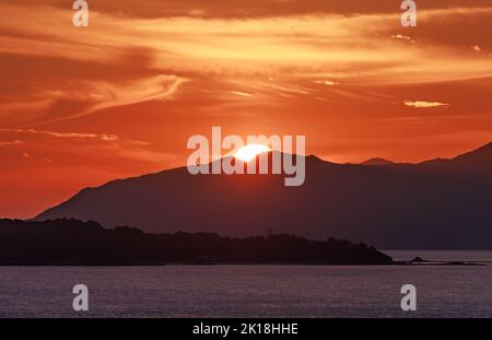 Die Sonne ragt über ferne Berge, während der Himmel in der Abenddämmerung orange leuchtet Stockfoto