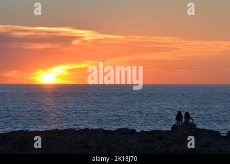 Sonnenuntergang in Cala Blanca, in der Nähe von Cuitadella, Menorca, mit Paaren, die gegen den Himmel geschildet sind Stockfoto