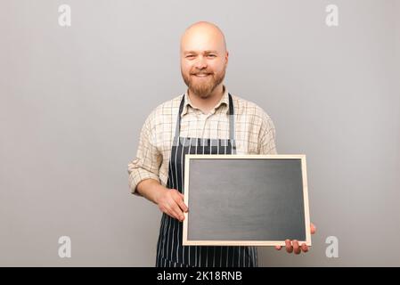 Ein kahlköpfiger Barista hält ein leeres, sauberes schwarzes Brett, um Platz für Kopien zu schaffen. Studio auf grauem Hintergrund. Stockfoto