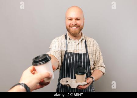 Ein junger kahler Mann reicht eine Tasse Kaffee zum Mitnehmen, während er eine andere hält. Studio auf grauem Hintergrund. Stockfoto