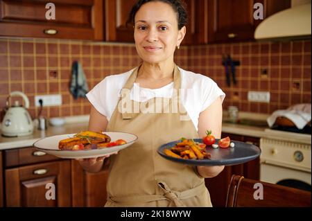 Charmante, multiethnische Frau auf der beigefarbenen Kochschürze trägt servierte Teller mit gesundem veganem Essen und gerösteten Süßkartoffeln Stockfoto