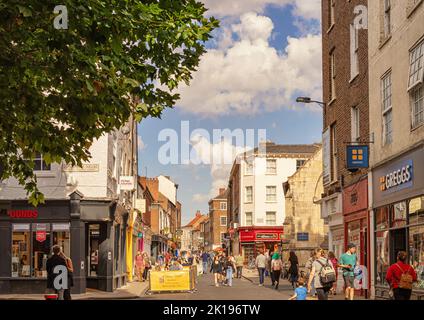 Eine Stadtstraße mit Geschäften und Restaurants im Sommer. Die Menschen gehen entlang und eine alte Kirche befindet sich auf der einen Seite. Ein Himmel mit Wolken ist oben. Stockfoto