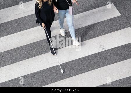 Die Szene einer blinden Frau, die auf einer Zebrakreuzung läuft, wurde von einer anderen Person mit ihrem weißen Stock unterstützt. Hilfe in den frühen Stadien der Blindheit Stockfoto