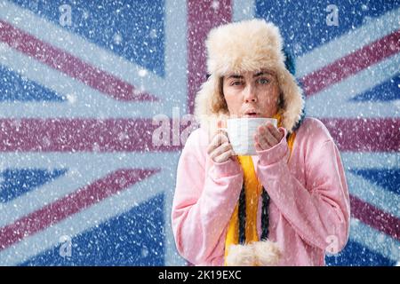 Energiekrise in Europa. Frau in einem Hut mit Ohrenklappen und einem Schal wärmt sich mit einer Tasse Tee. Im Hintergrund ist die Flagge von Großbritannien und ein Schnee Stockfoto