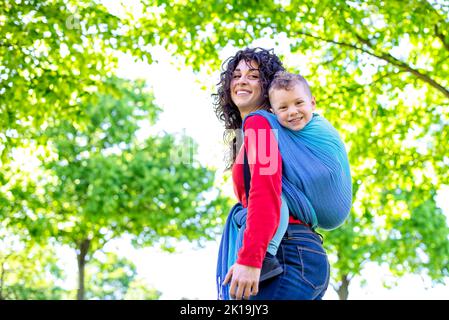 Die glückliche junge Mutter trägt ein Kind auf dem Rücken, das in einem öffentlichen Parkgarten mit einem bunten traditionellen Stoffstück zusammengebunden ist. Mutter und Sohn in einem Baby wra Stockfoto