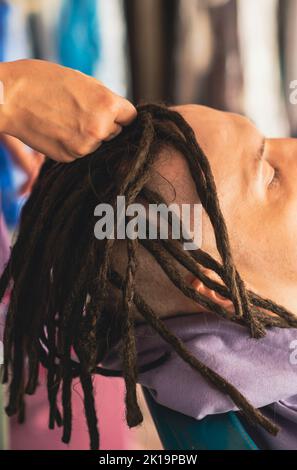 Eine nicht erkennbare Person flechtiert Dreadlocks für den Mann im Salon. Gesichtslose professionelle Friseur macht Frisur für Kunden. Nahaufnahme Stockfoto