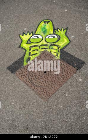 Europa, Frankreich, Dunkerque - 9. Juli 2022: Nahaufnahme. Kunstvoll bemalter grüner Kopf von Krokodilkrabbeln unter brauner Metall-Kanalabdeckung. Stockfoto