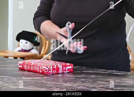 Womans Hand schneidet mit einer Schere ein Band eines eingewickelten Weihnachts- oder anderen Weihnachtsgeschenks. Rot-weiß Verpackung, Band und Schere im DIY Hobby. Stockfoto