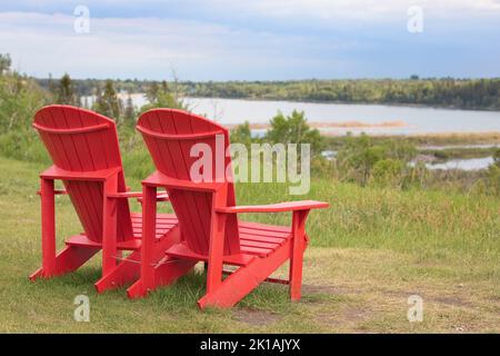 Rote Adirondack-Stühle mit Blick auf einen See in Weaselhead Flats, einem Naturgebiet in Calgary, Alberta, Kanada Stockfoto