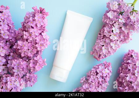 Mockup weiße Squeeze-Flasche Kosmetikschlauch und lila Blüten auf blauem Hintergrund. Creme, Gleitgel, Feuchtigkeitscreme, Hautpflege, Sonnencreme. Flach liegend, oben Stockfoto