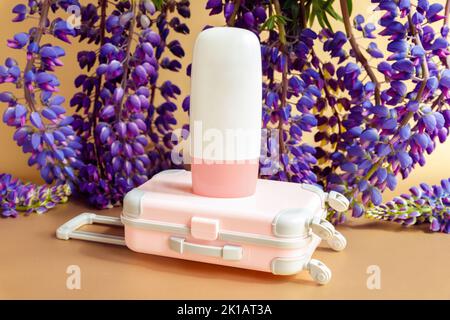 Mockup weiße Kosmetikröhrchen-Flasche mit rosa Schraubverschluss, kleinem rosa Reisekoffer-Spielzeug und lila Lupine-Blüten viel auf braunem Hintergrund. Mockup, fr Stockfoto