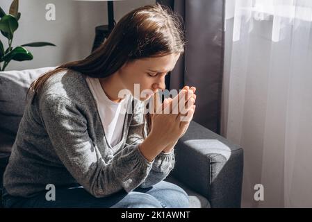 Nahaufnahme einer ängstlich jungen Frau, die auf einem grauen Sofa sitzt und mit gefalteten Händen Gott betet, die eine Lebenskrise hat. Nervöse Brünette Frau 20s versucht, konzentra Stockfoto