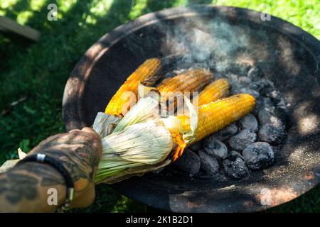 Braten von Maiskolben auf heissem Kohlegrill, Person Hand mit Tattoo Stockfoto