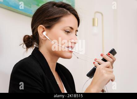 Seitenansicht der jungen weiblichen Mitarbeiterin im formellen Anzug mit Kopfhörer sprechen auf dem Mobiltelefon, während Kunden während der Telearbeit beraten Vom Heimbüro Stockfoto