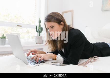 Seitenansicht der jungen weiblichen Remote-Spezialist in formelle Kleidung Liegen auf dem Bett und arbeiten online auf Laptop in Licht Schlafzimmer zu Hause Stockfoto