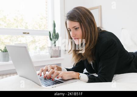 Seitenansicht der jungen weiblichen Remote-Spezialist in formelle Kleidung Liegen auf dem Bett und arbeiten online auf Laptop in Licht Schlafzimmer zu Hause Stockfoto