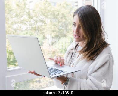 Junge Frau im weißen Bademantel, die auf dem Laptop im Internet surfen kann, während sie am sonnigen Morgen zu Hause in der Nähe des Fensters steht Stockfoto