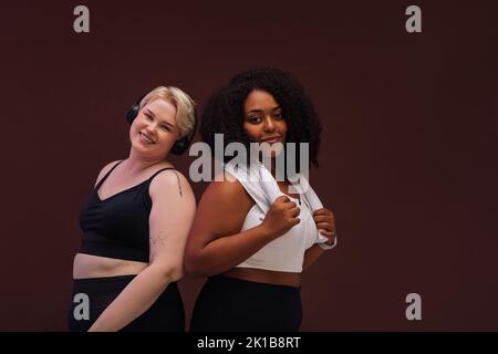 Zwei plus size Frau, die vor braunem Hintergrund Rücken an Rücken steht und im Studio auf die Kamera schaut Stockfoto