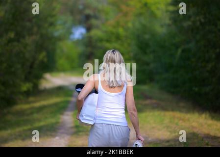 Rückansicht einer attraktiven blonden Frau mit einer Tasche und einem Badetuch unter ihrem Arm auf dem Weg zum oder vom See Stockfoto