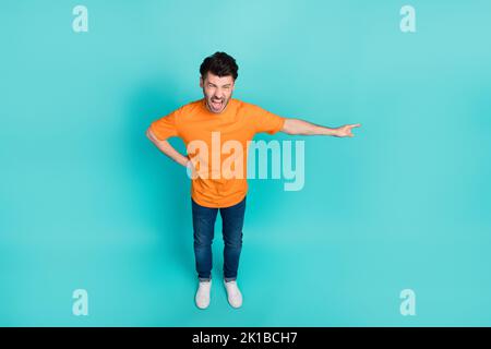 In voller Länge Foto von jungen lustig gut aussehend agressive Mann tragen orange T-Shirt Denim Jeans Finger zeigt leeren Raum wütend Chef isoliert auf cyan Farbe Stockfoto
