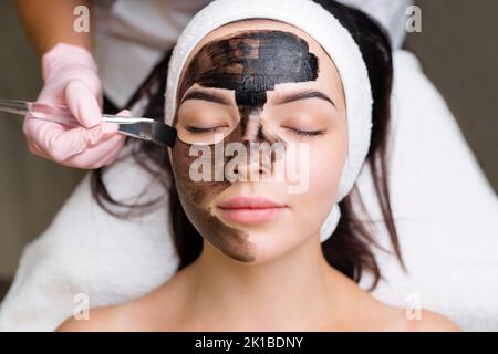 Kosmetologe, der schwarze Maske auf hübsches Frauengesicht mit schwarzen Handschuhen aufträgt, wunderschöne Frau im Spa, die Gesichtsbehandlungen hat Stockfoto