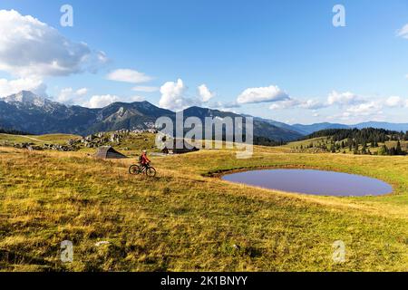 Frau Touristin auf einem Mountainbike, Radfahren auf einer Velika planina Almwiese, mit hölzernen Hirtenhütten und einem Teich auf der Rückseite, kamnik, Slowenien Stockfoto