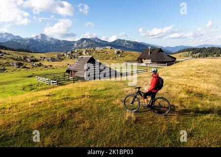 Frau Touristin auf einem Mountainbike, Radfahren auf einer Almwiese von Velika planina, mit traditionellen Holzhütten auf der Rückseite, Kamnik, Slowenien Stockfoto