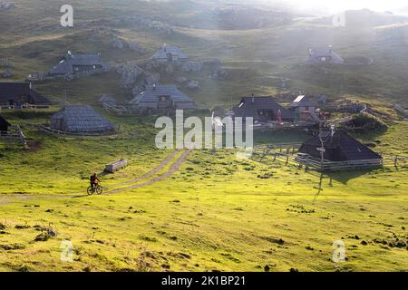 Ein Mountainbiker auf dem Velika Planina Plateau in der Nähe von Kamnik, bei Sonnenuntergang zu traditionellen Hirtenhäusern aus Holz, Slowenien Stockfoto