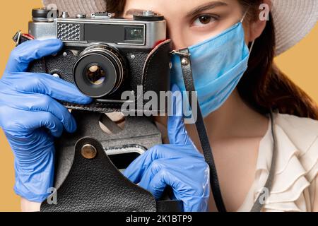 Foto aufnehmen. Covid-19-Schutz. Halten Sie Momente fest. Urlaubsreise. Frau in Medizin Maske Handschuhe fotografieren auf Retro-Kamera isoliert auf beige. Stockfoto