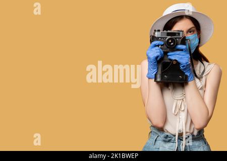 Foto aufnehmen. Covid-19-Schutz. Halten Sie Momente fest. Urlaubsreise. Frau in Medizin Maske Handschuhe fotografieren auf Retro-Kamera isoliert auf beige. Stockfoto