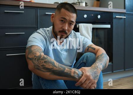 Der junge asiatische Mann sitzt auf dem Boden in der Küche und umarmt seine Knie Stockfoto