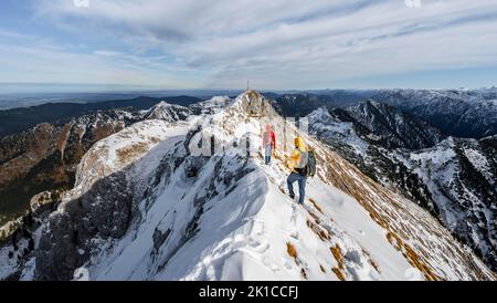 Zwei Bergsteiger auf einem schmalen, felsigen, schneebedeckten Bergrücken, im hinteren Gipfel der Ammergauer Hochplatte mit Gipfelkreuz, Blick auf das Bergpanorama, Wandern Stockfoto