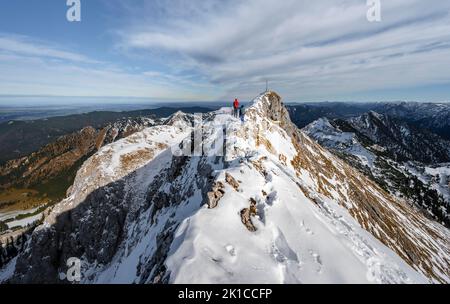 Zwei Bergsteiger auf einem schmalen, felsigen, schneebedeckten Bergrücken, im hinteren Gipfel der Ammergauer Hochplatte mit Gipfelkreuz, Blick auf das Bergpanorama, Wandern Stockfoto