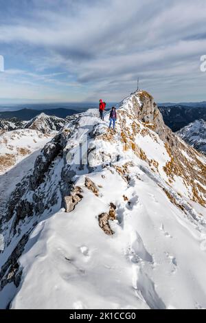 Zwei Bergsteiger auf einem schmalen, felsigen, schneebedeckten Bergrücken, im hinteren Gipfel der Ammergauer Hochplatte mit Gipfelkreuz, Blick auf das Bergpanorama, Wandern Stockfoto