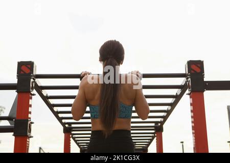 In der Rückansicht zeigt sich eine athletische Frau, die auf einer horizontalen Leiter im Freien Pull-Ups macht Stockfoto