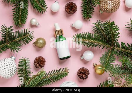 Grüne Tropfflasche in der Nähe von Weihnachtsdekorationen, Tannenzweigen und Tannenzapfen auf rosa Draufsicht. Verpackungsmockup der Marke Holiday. Hautpflegeprodukt Stockfoto