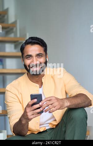 Glücklicher indischer Mann, der zu Hause in Ohrhörern sitzt und das Telefon senkrecht hält. Stockfoto